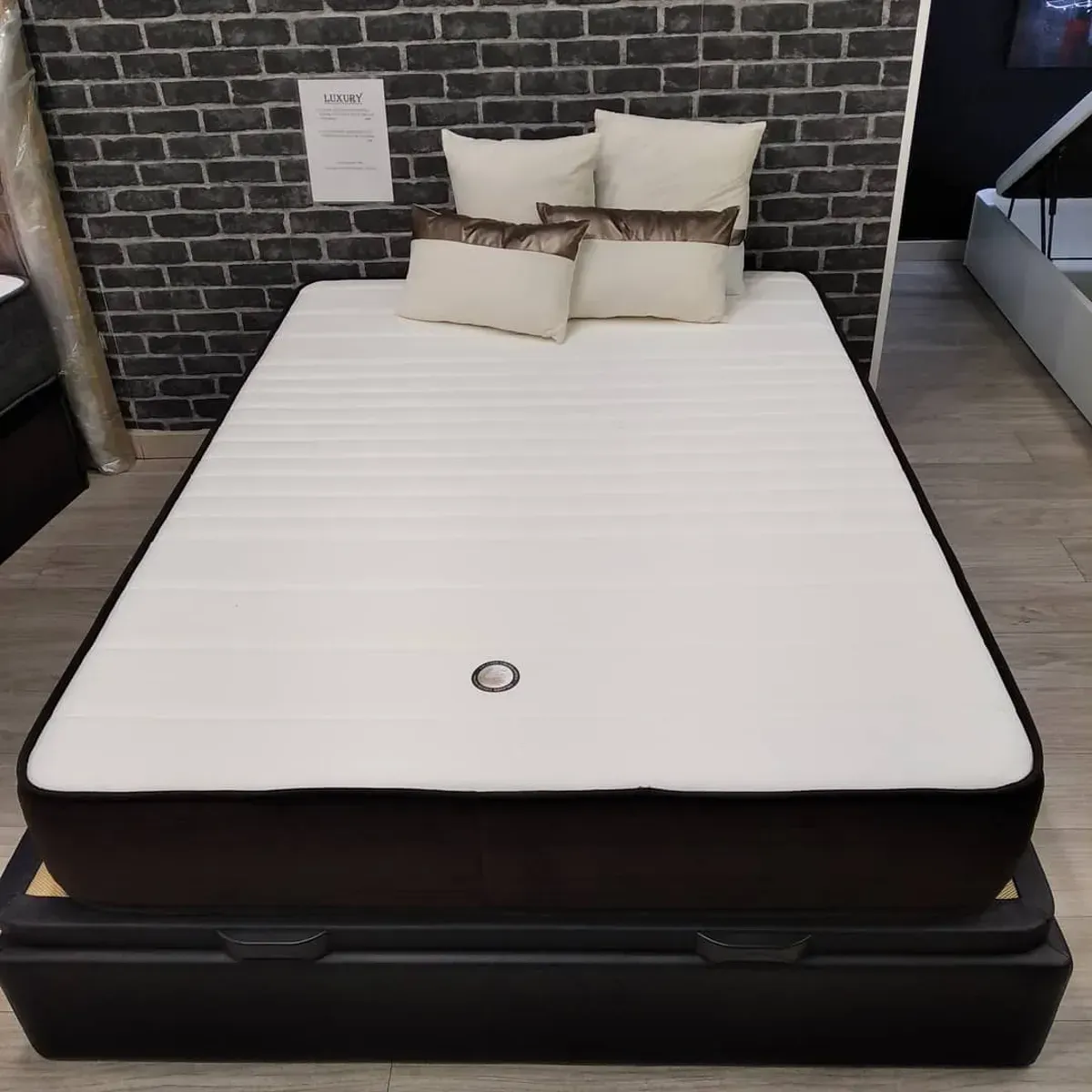 Una cama con colchón blanco, ribete marrón oscuro y varias almohadas, sobre una base negra.