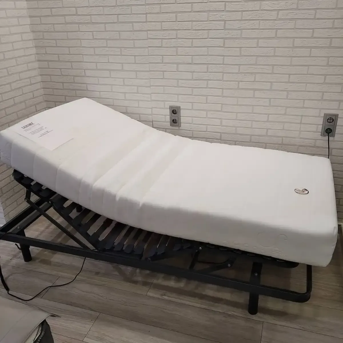 Una estructura de cama blanca ajustable con un colchón blanco está apoyada contra una pared de ladrillo.