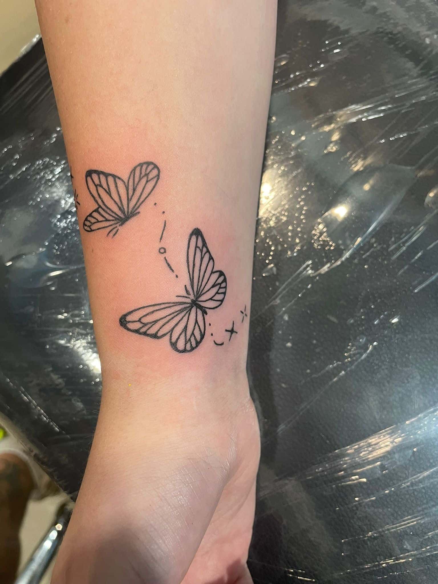 Dos tatuajes de mariposas en la muñeca, una de ellas en vuelo. Contorno negro.