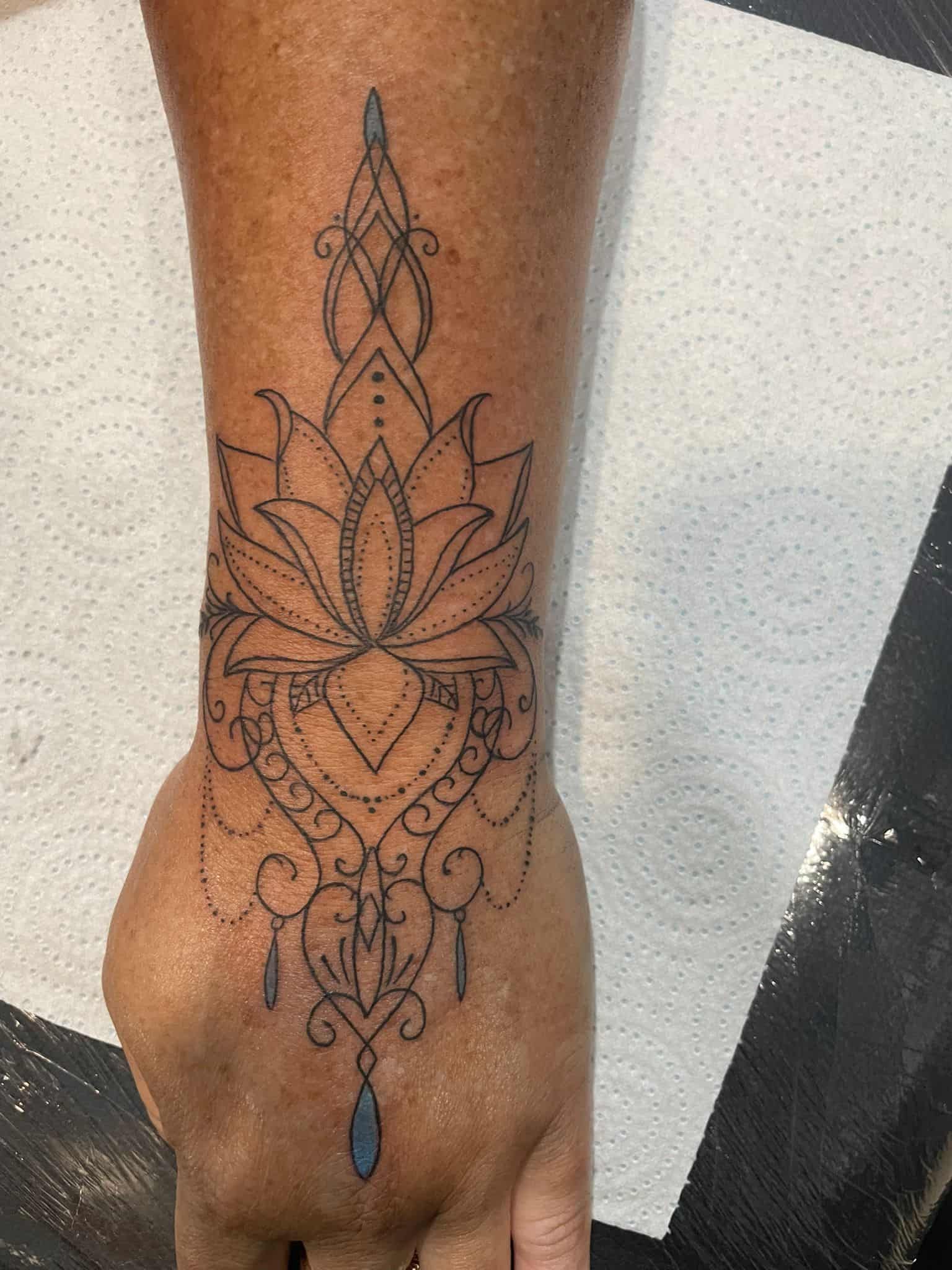 Tatuaje en la muñeca: diseño de flor de loto adornado con gota azul y líneas negras detalladas.