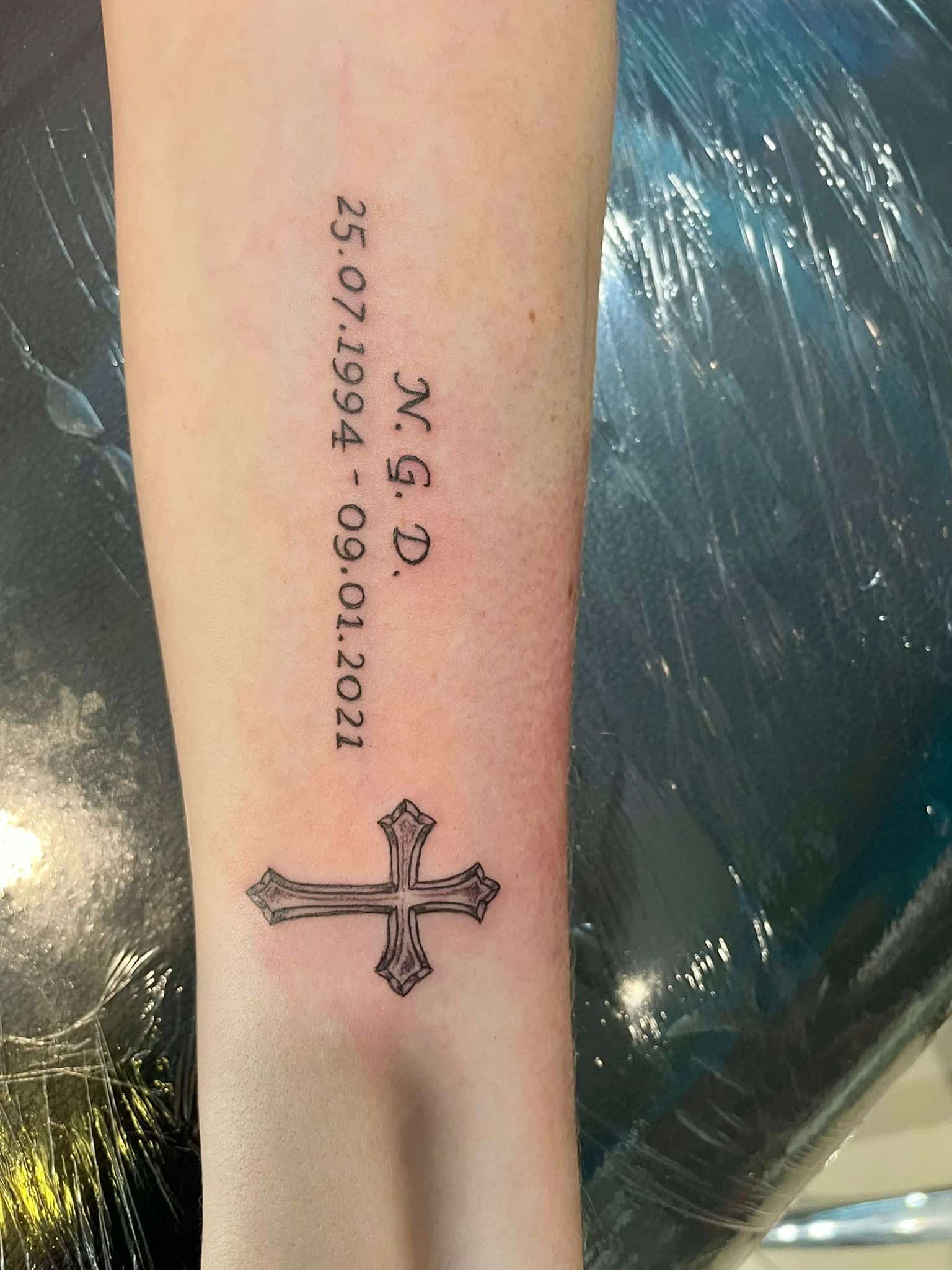 Tatuaje de una cruz y fechas: 25.07.1994 - 09.01.2021, y las iniciales