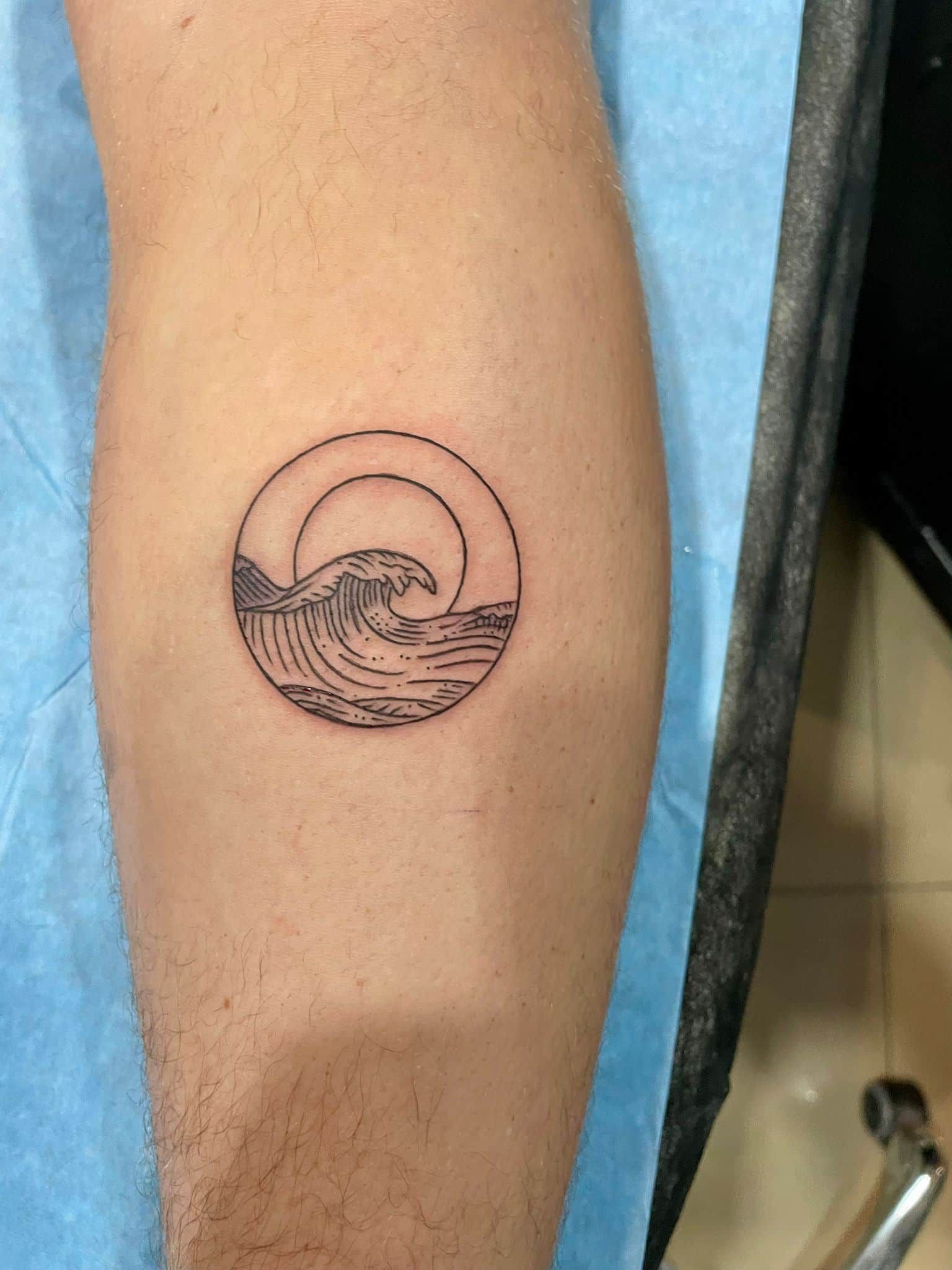 Tatuaje en tinta negra de una ola del océano que se extiende dentro de un círculo, en la pierna de una persona.