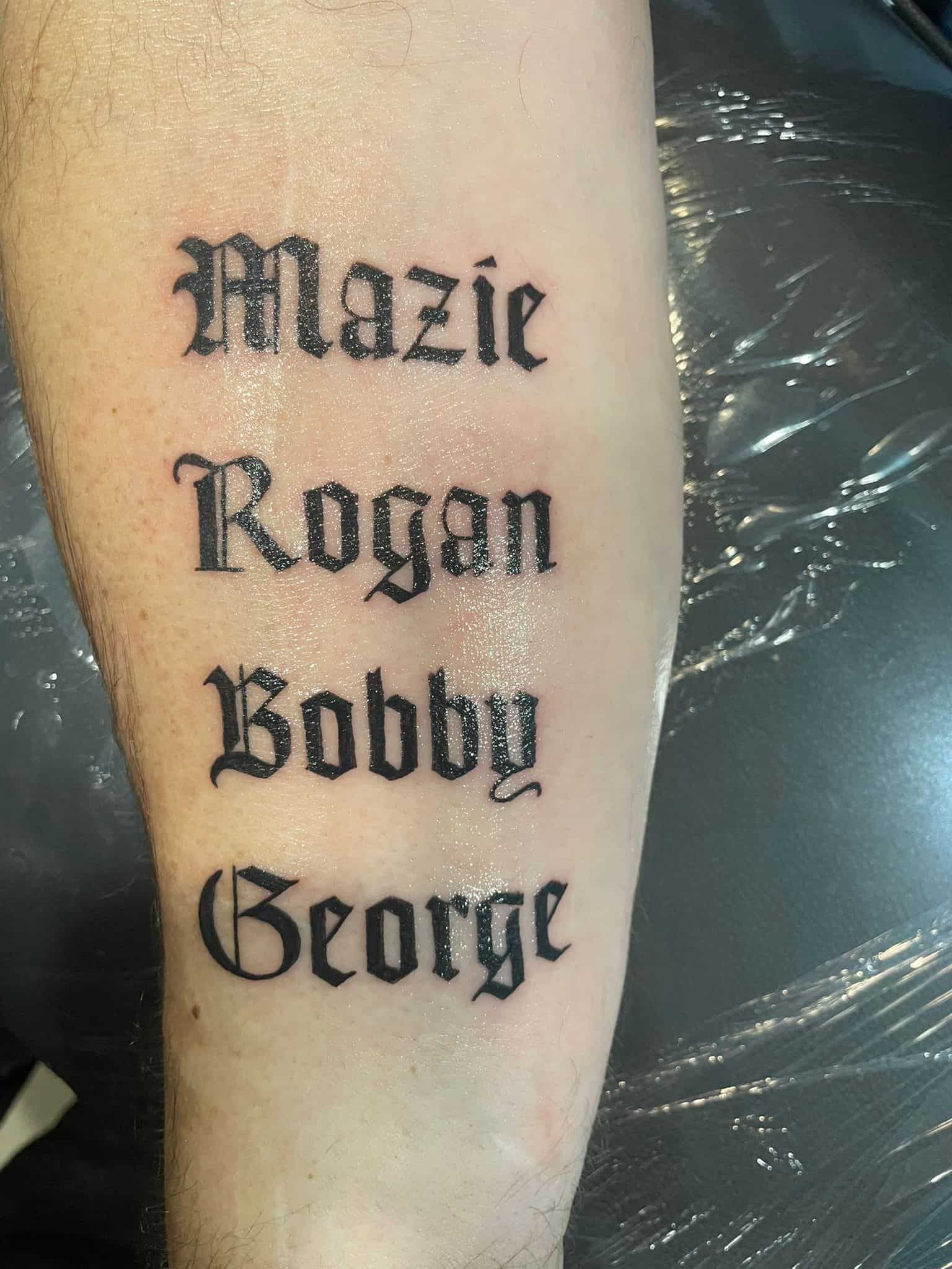 Tatuaje en el brazo con cuatro nombres: Mazie, Rogan, Bobby, George, en escritura gótica.
