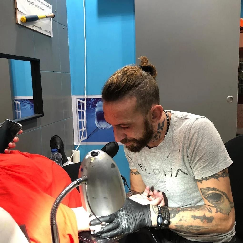 Hombre con tatuajes trabajando en un tatuaje, con guantes y una camisa gris en una tienda.