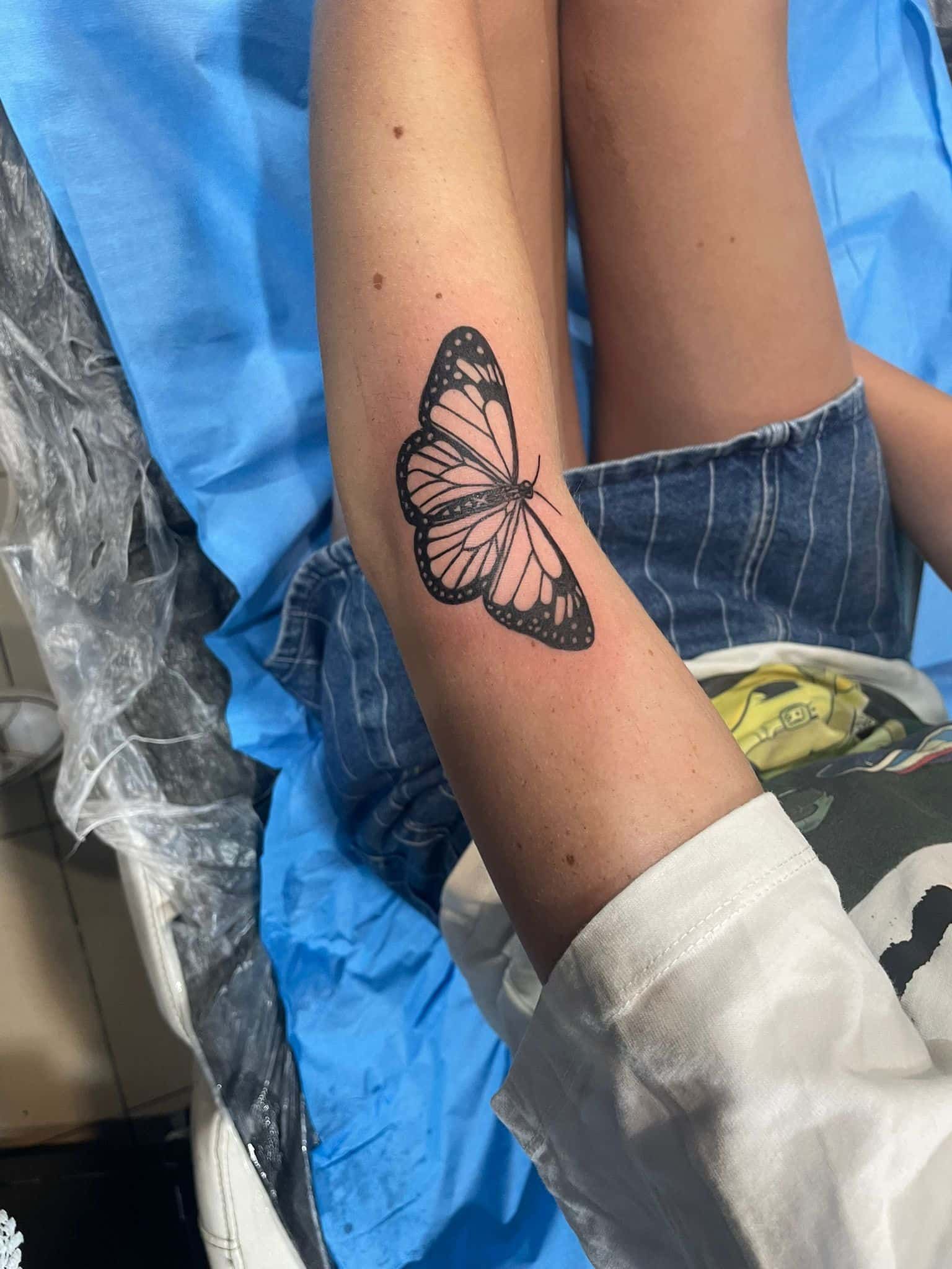 Tatuaje de mariposa negra en la parte interna del brazo. Lleva pantalones cortos de mezclilla y camisa blanca.