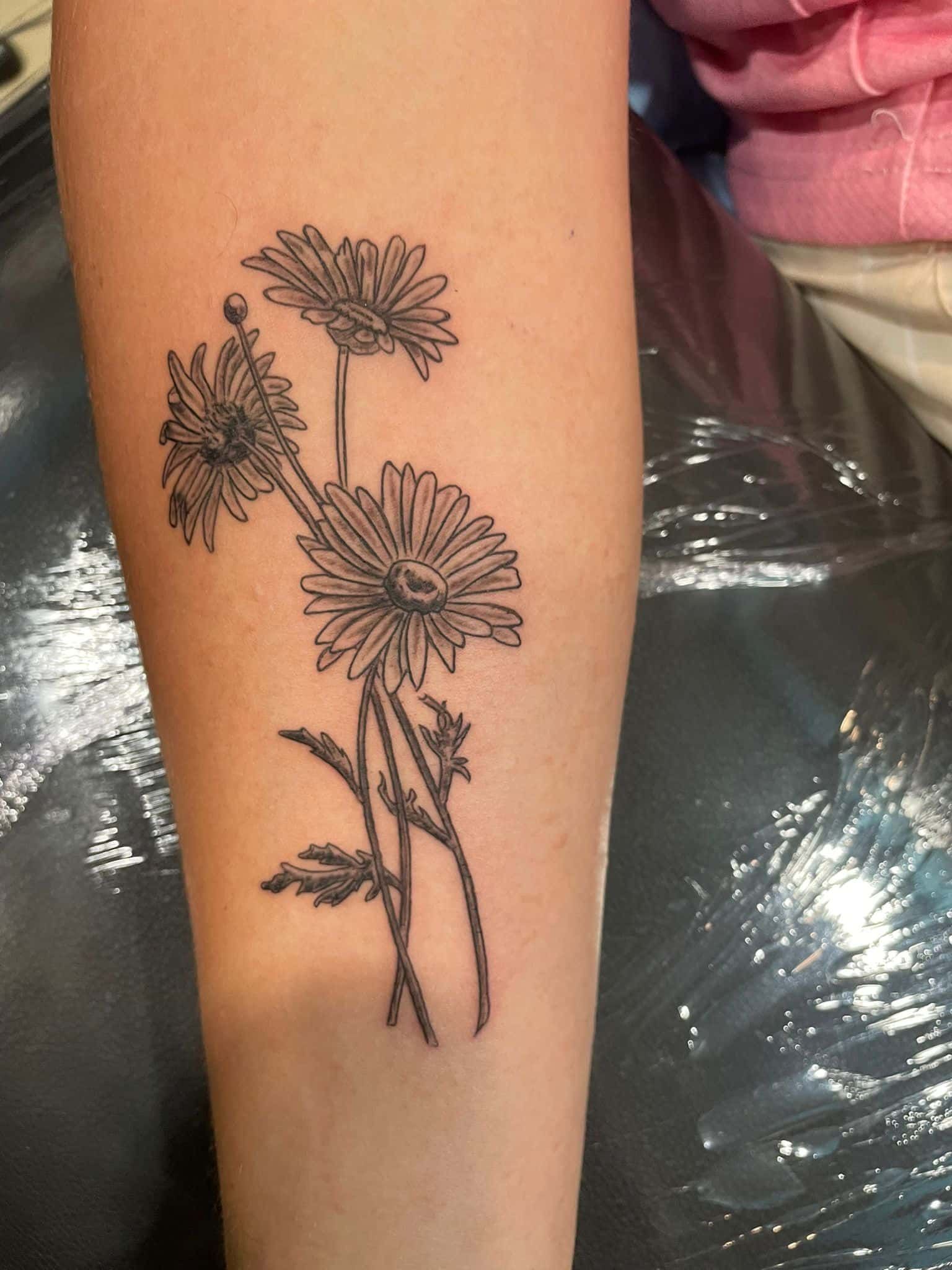 Tatuaje de margarita en tinta negra en el antebrazo, con pétalos y tallos detallados.
