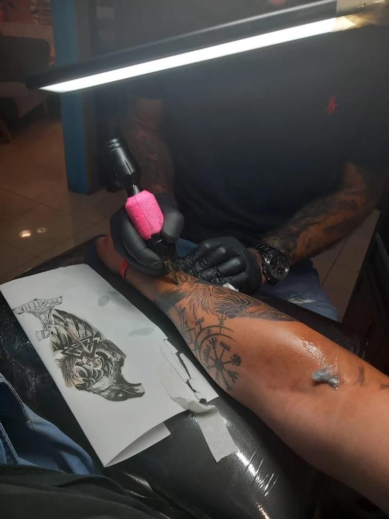 Artista del tatuaje con guantes negros, utilizando una máquina en la pierna de una persona, con una plantilla de diseño.