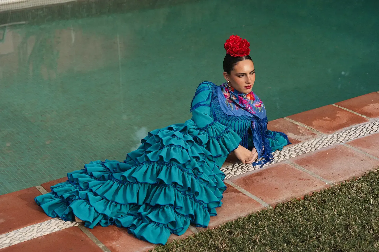 Mujer con vestido de flamenca color verde azulado junto a la piscina, con chal y flor en el pelo.