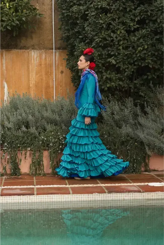 Mujer con vestido de flamenca con volantes color verde azulado y pañuelo, caminando junto a una piscina. Flores rojas en el pelo.