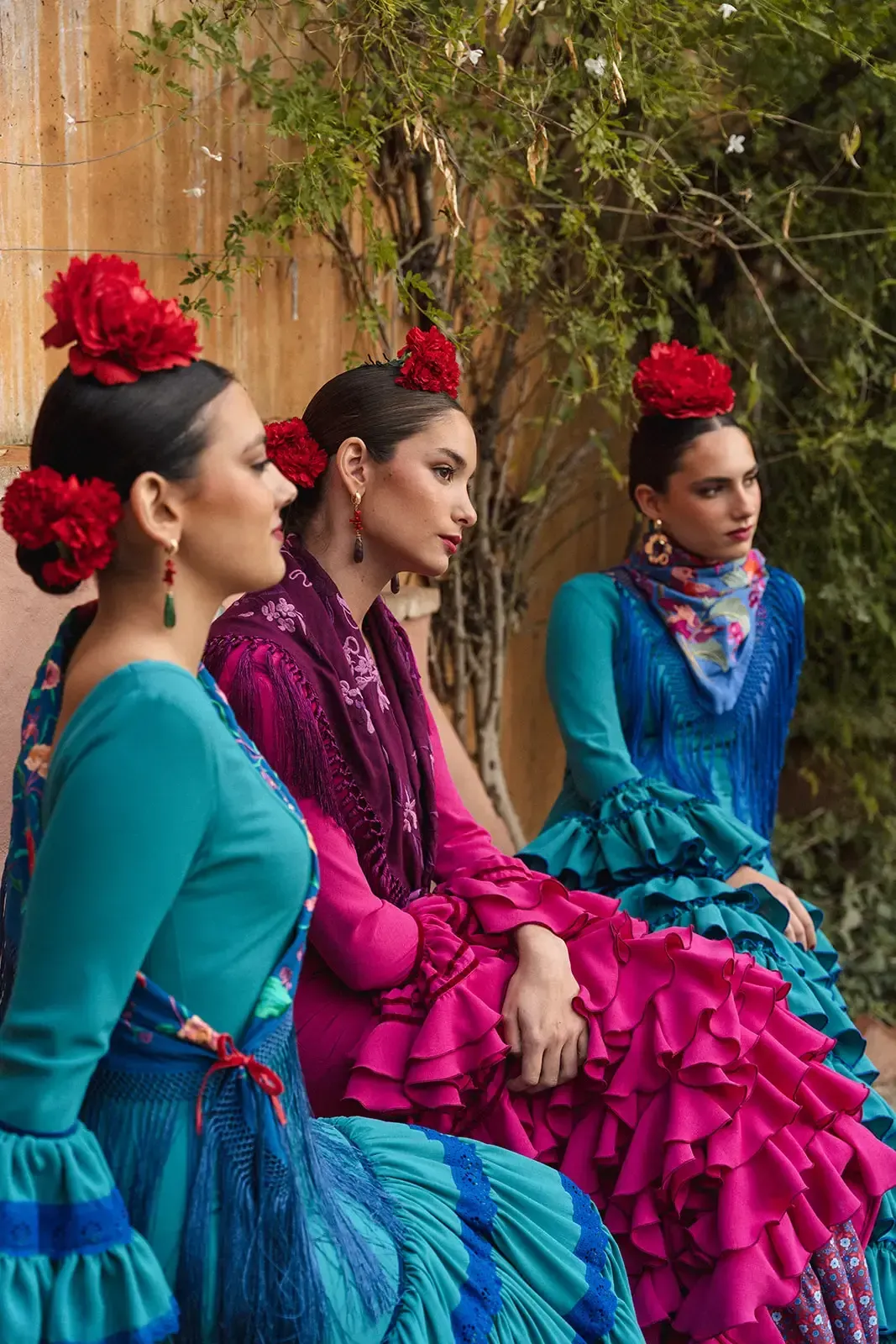Tres mujeres con vibrantes trajes de flamenca posando al aire libre. Cada una luce un tocado floral y faldas con volantes.