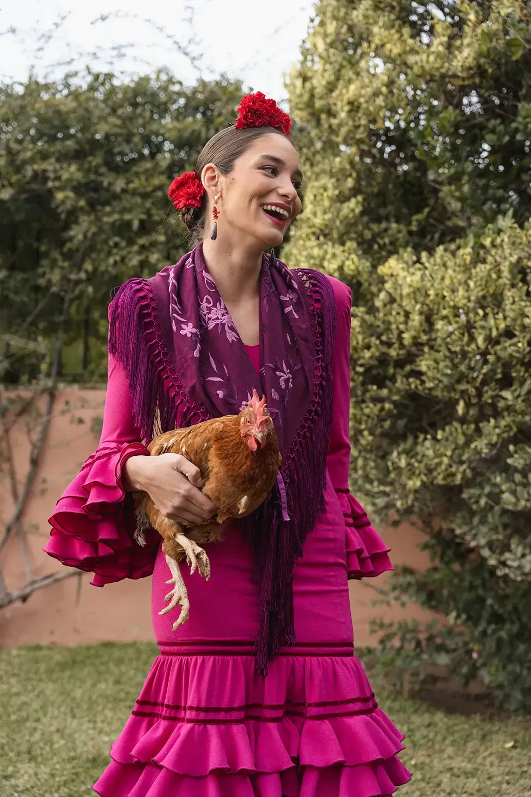 Mujer con traje de flamenca fucsia sosteniendo un pollo marrón, sonriendo, en un jardín.
