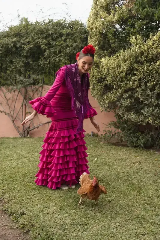 Mujer con vestido de flamenca color magenta con un pollo en un patio con césped.