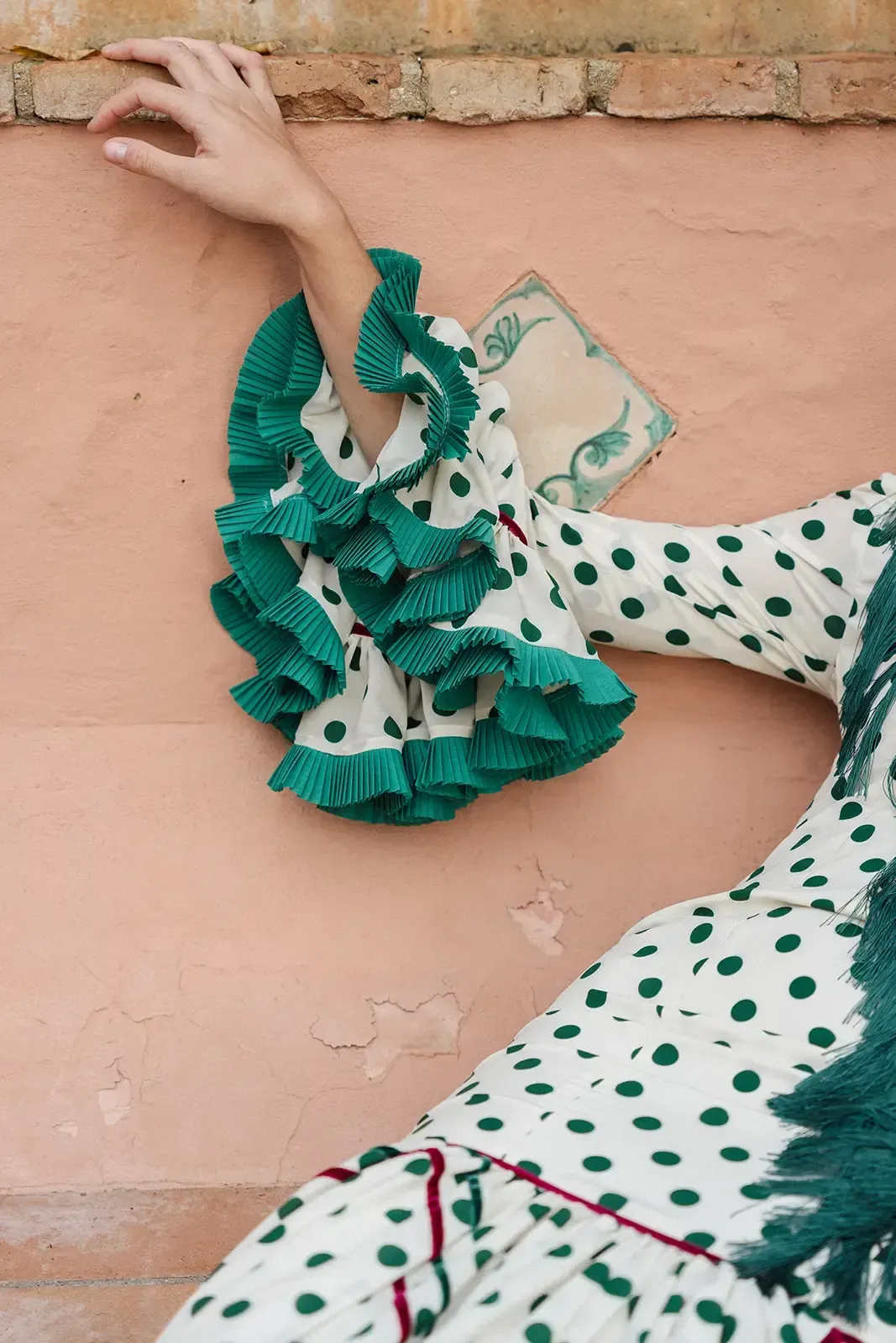 Brazo de mujer con un vestido de lunares con mangas verdes con volantes, apoyado contra una pared rosa.