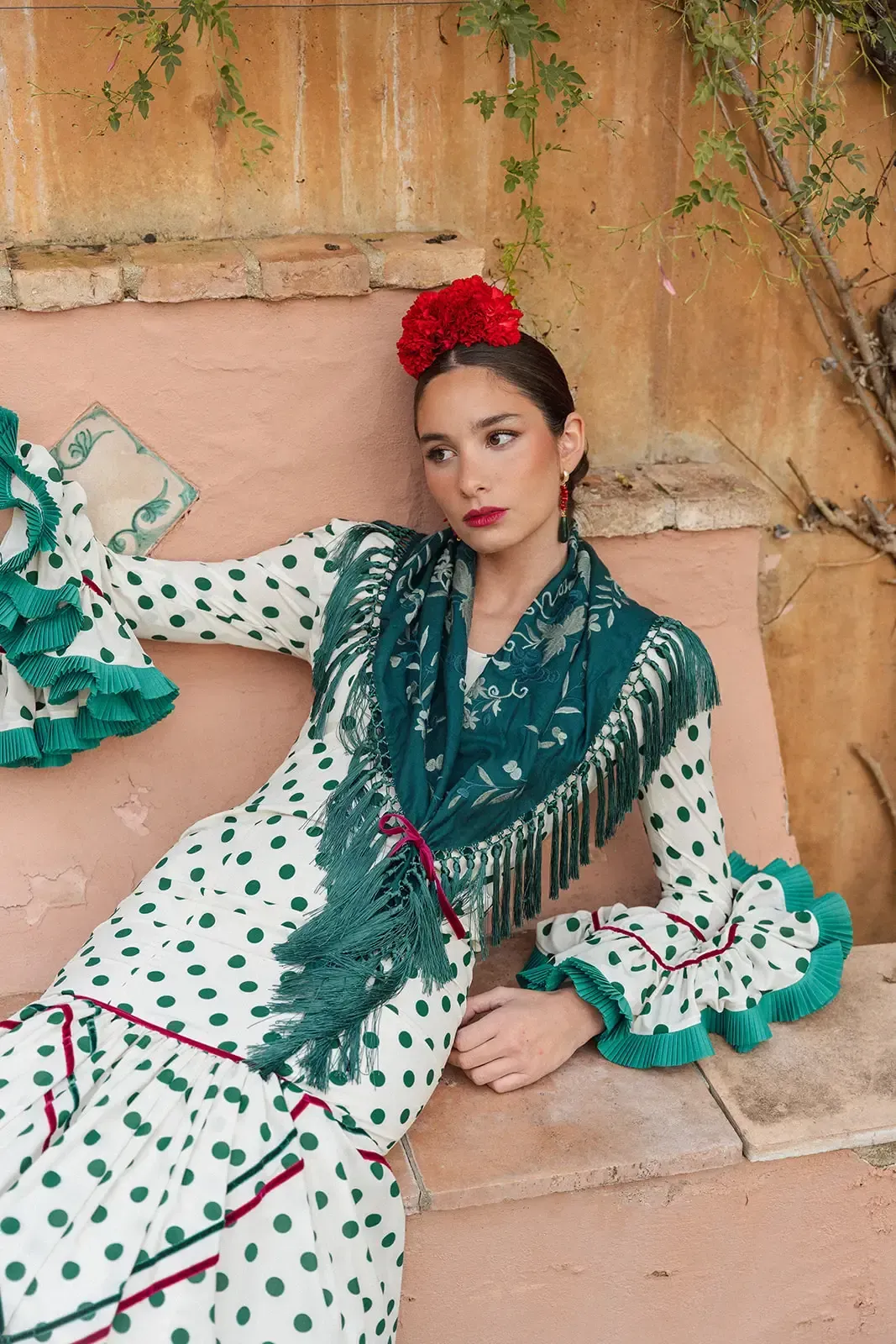 Mujer con traje de flamenca con lunares, mantón verde y flor roja, sentada al aire libre.
