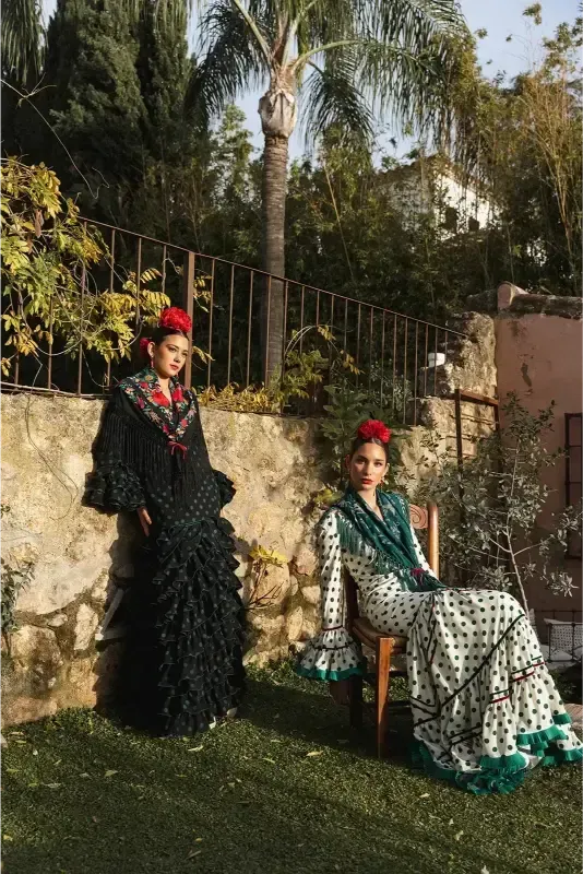 Dos mujeres con trajes de flamenca posan en un jardín, una de pie y otra sentada; llevan tocados de flores.