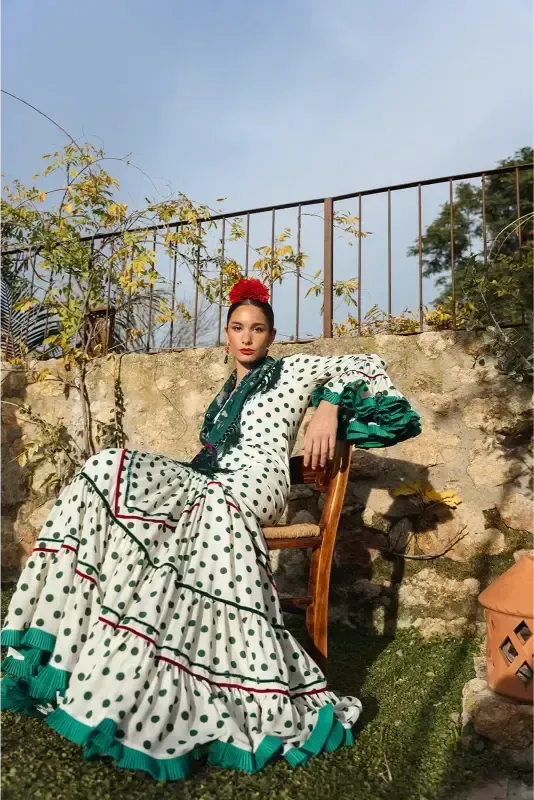 Una mujer con un vestido blanco de lunares con ribete verde se sienta en una silla al aire libre y tiene una flor roja en el pelo.