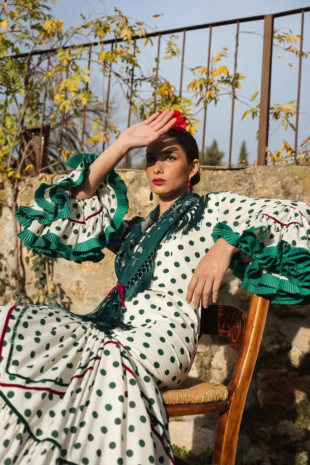Mujer con traje de flamenca blanco de lunares y ribete verde, protegiéndose los ojos. Sentada en una silla al aire libre.