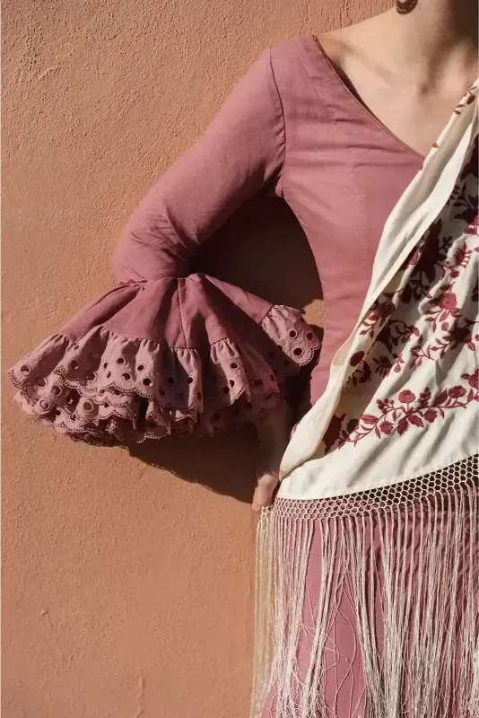 Vestido rosa con mangas acampanadas con ojales, chal crema con flecos, contra una pared color melocotón.