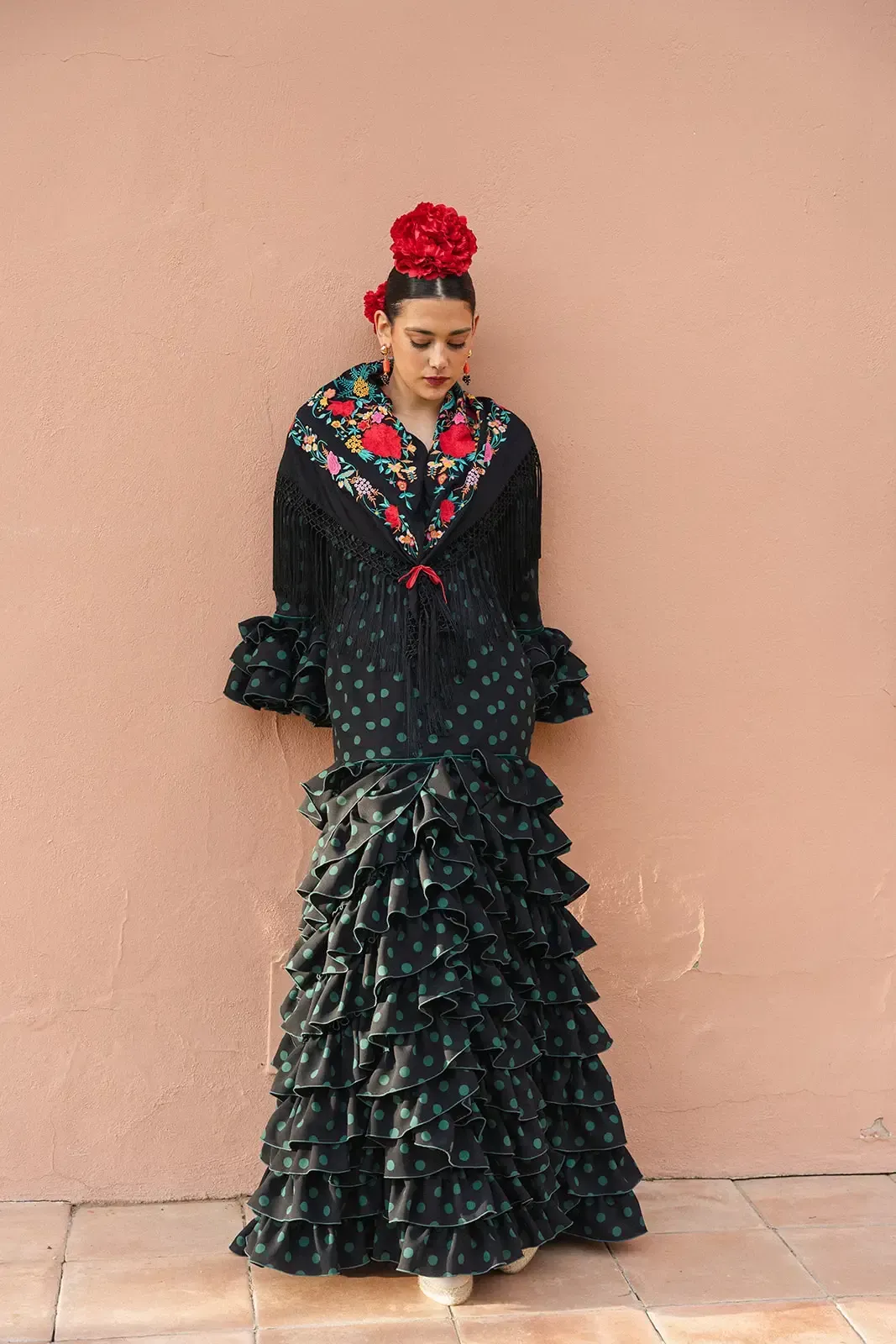 Mujer con traje de flamenca con volantes, apoyada contra una pared rosa, flor roja en el pelo.
