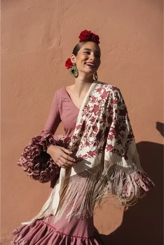 Mujer con traje de flamenca y mantón, sonriendo, contra pared rosa.