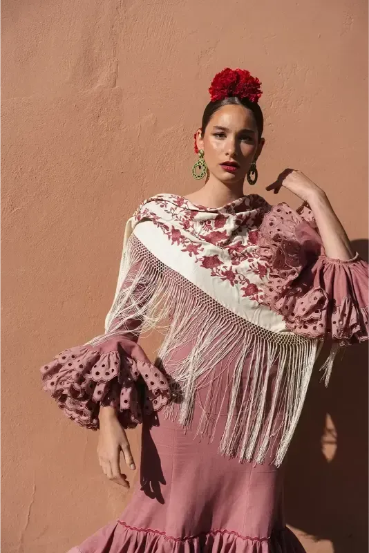 Mujer con vestido de flamenca rosa y flor en el pelo, de pie frente a una pared color melocotón.