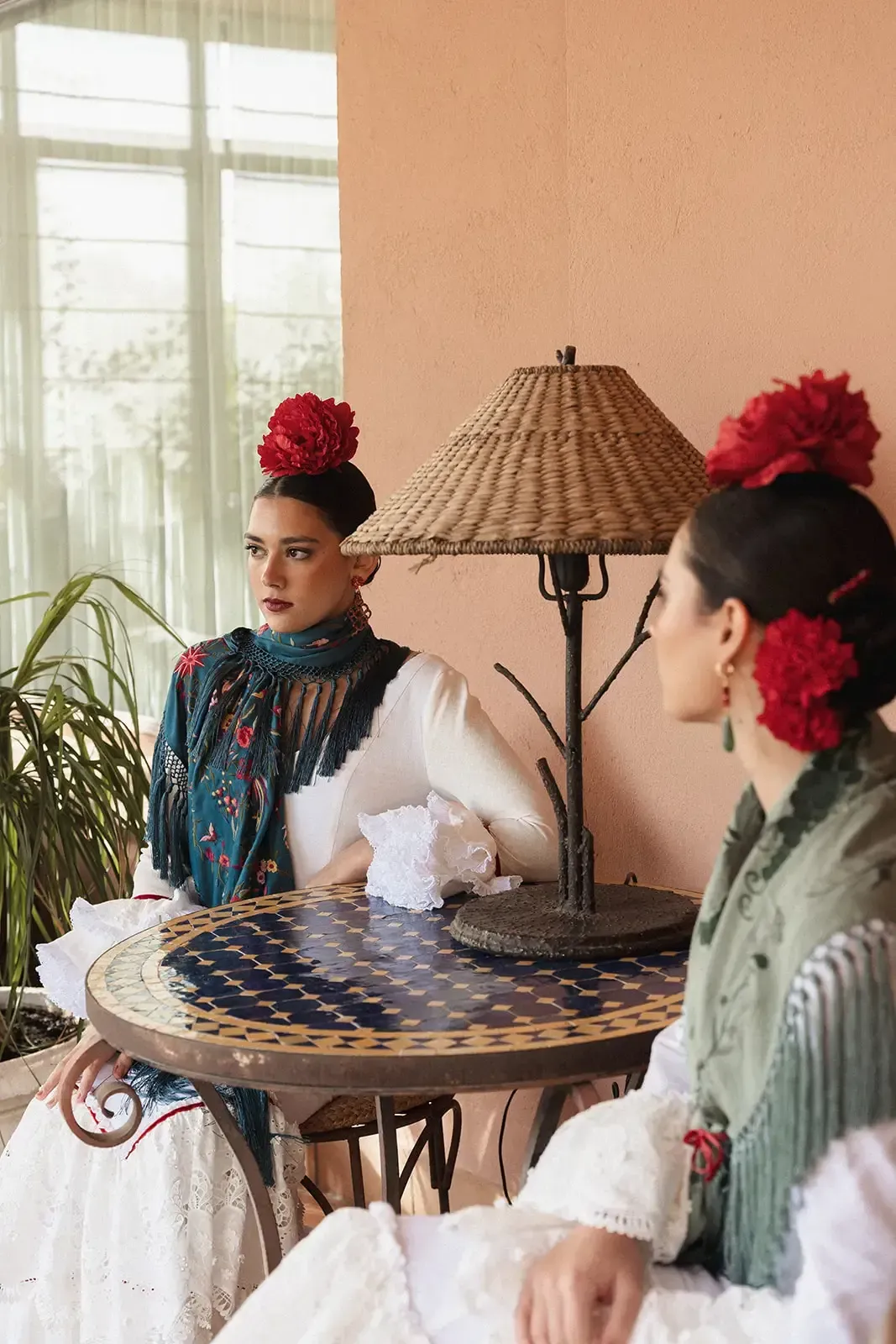 Dos mujeres con atuendos tradicionales están sentadas a una mesa al aire libre. Llevan adornos florales rojos para el cabello.