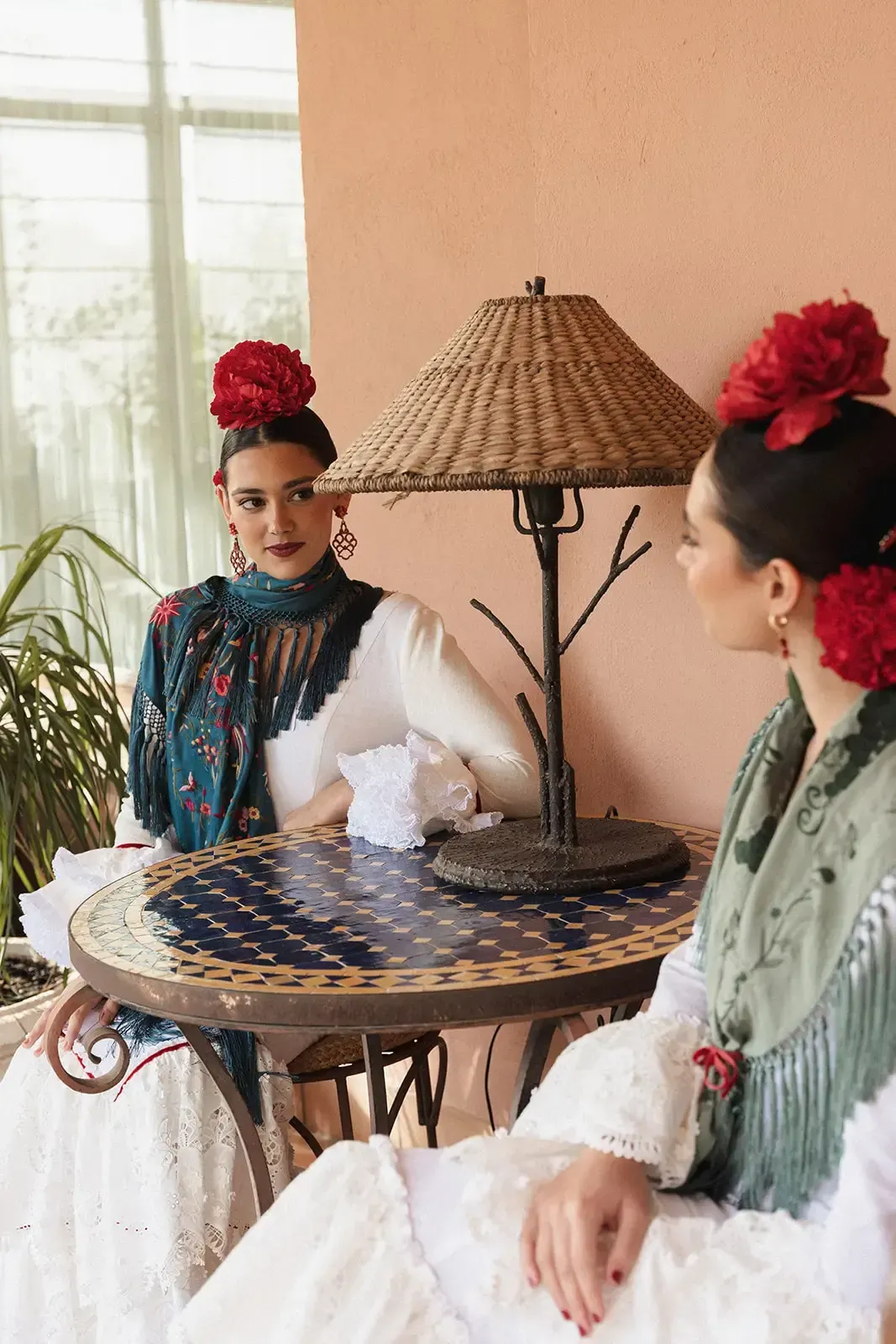 Dos mujeres con vestidos blancos tradicionales, tocados con flores rojas y chales estampados se sientan en una mesa bajo una lámpara.