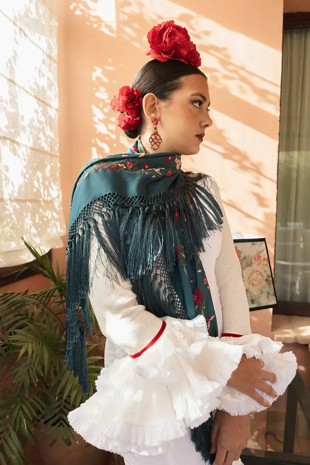 Mujer con vestido blanco con mangas con volantes, chal verde azulado, flores rojas en el pelo, mirando hacia otro lado.