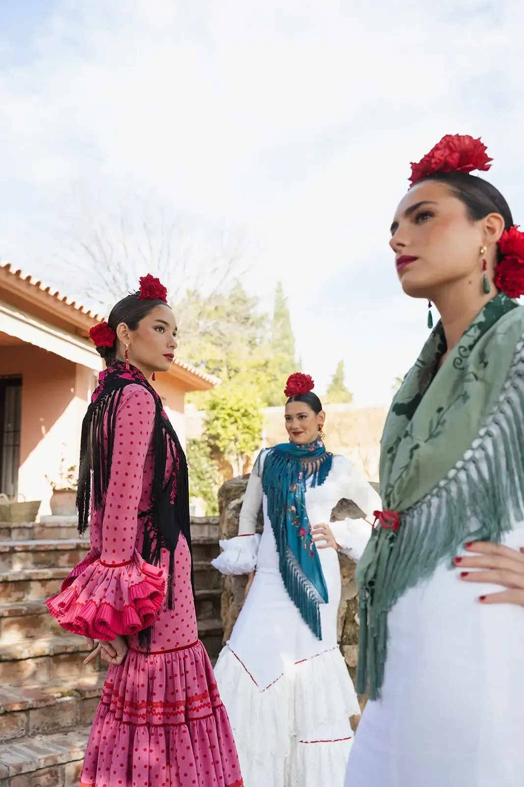 Tres mujeres con trajes de flamenca posando al aire libre. Cada una lleva una flor en el pelo y un chal de colores rosa, blanco y verde azulado.