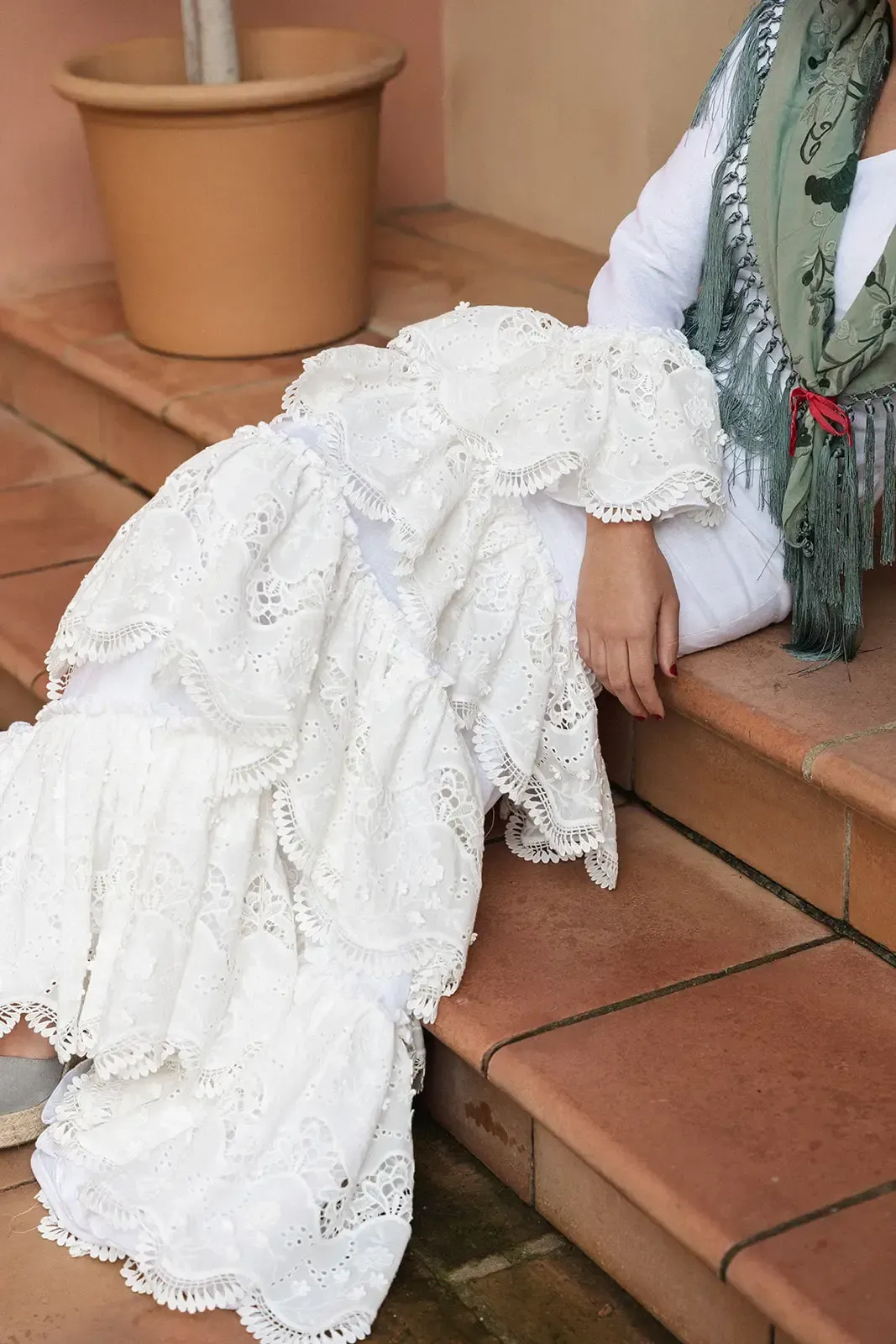 Una mujer con un vestido blanco con ojales se sienta en unos escalones de terracota y lleva un chal verde.