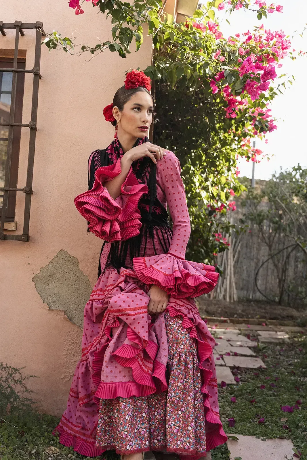 Mujer con traje de flamenca rosa con volantes, posando junto a una pared rosa y flores.