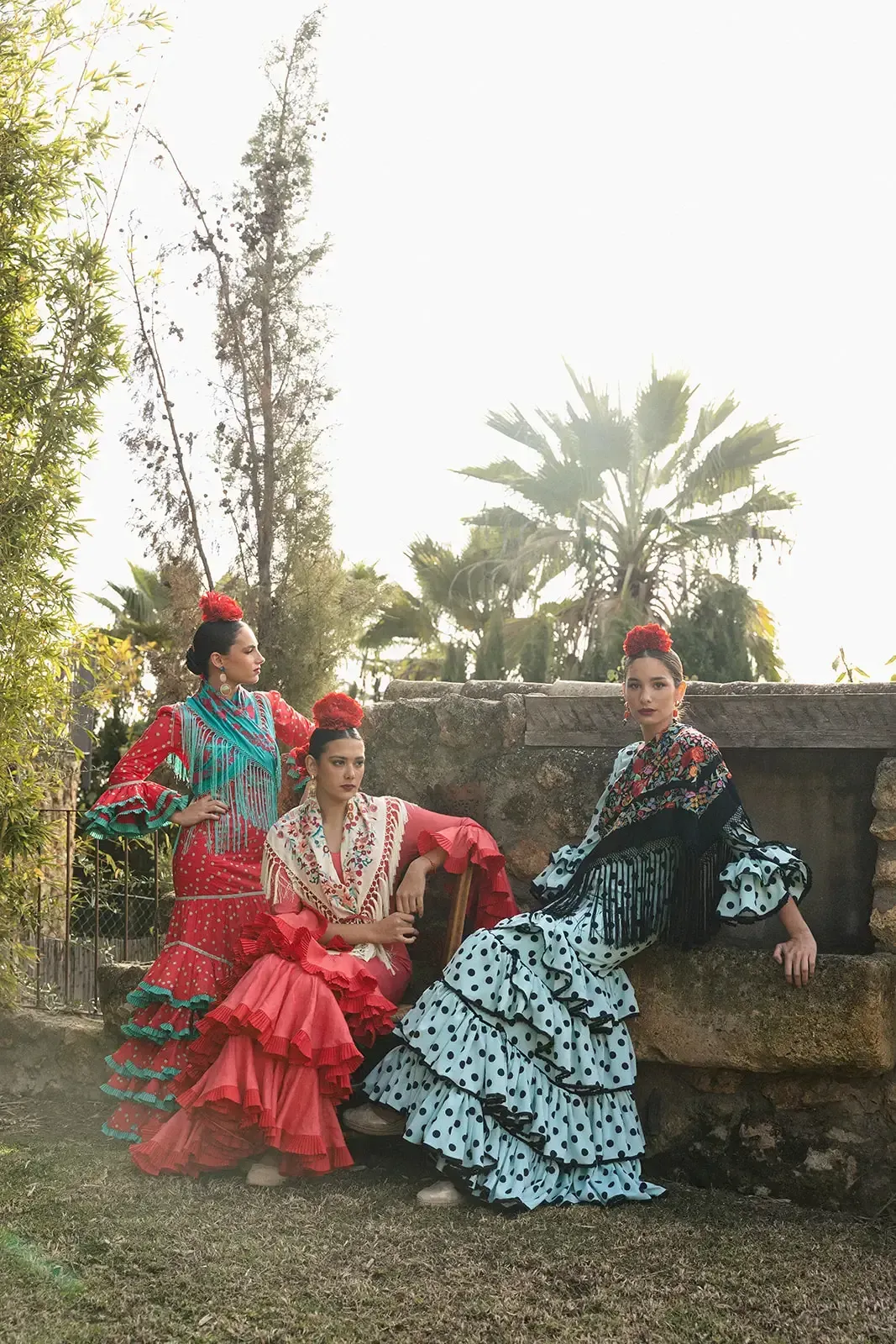 Tres mujeres con trajes de flamenca posan al aire libre; vestidos rojos, verde azulado y negros, tocados florales.