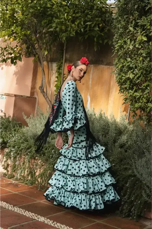 Mujer con un vestido de flamenca de lunares verde azulado y una flor roja, de pie al aire libre, sosteniendo un chal negro.