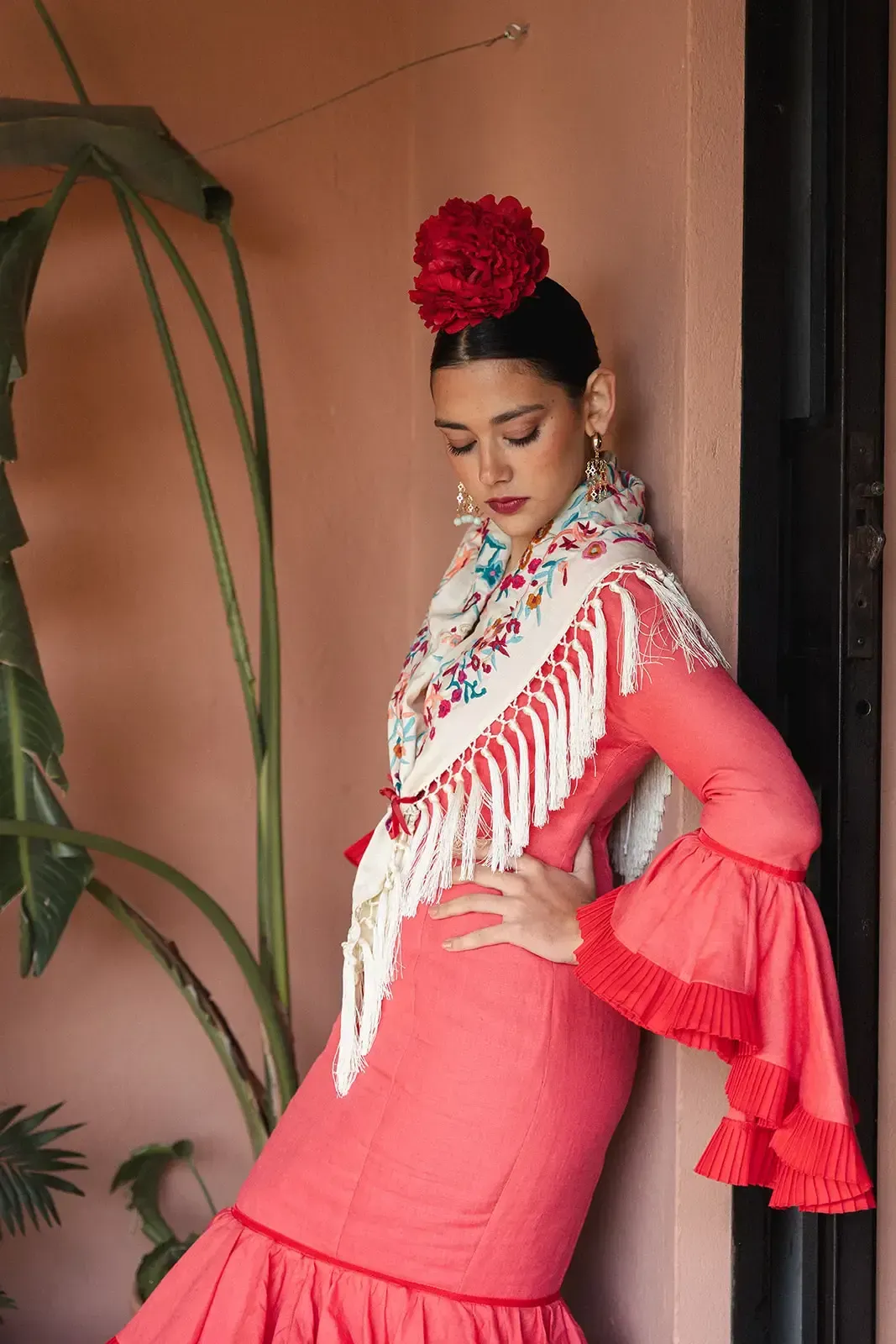 Mujer con un traje de flamenca coral y una flor roja en el pelo, apoyada contra una pared rosa.