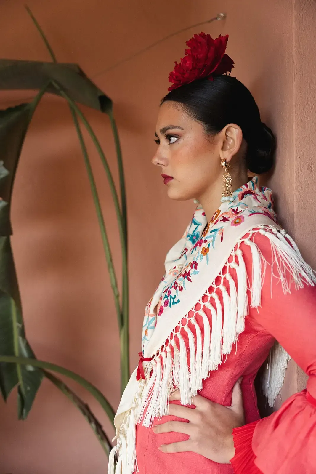 Mujer con vestido rojo y chal con flecos, flor roja en el pelo, apoyada contra una pared rosa.
