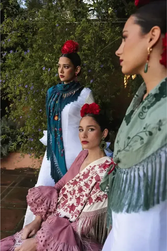 Tres mujeres con trajes de flamenca posando al aire libre. Cada una lleva una flor en el pelo y un mantón.