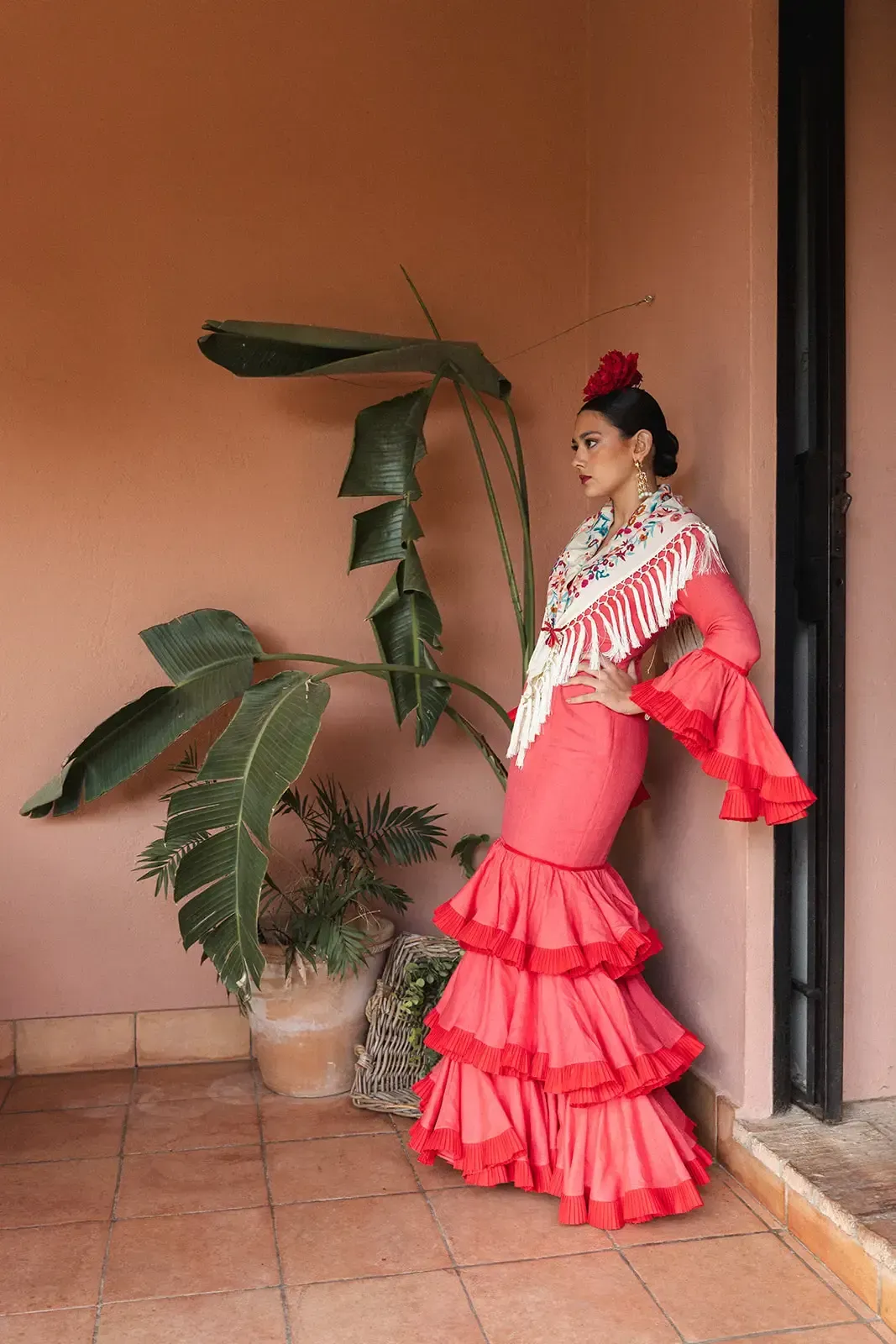 Mujer con vestido de flamenca rojo se apoya en una pared de terracota con una gran planta.