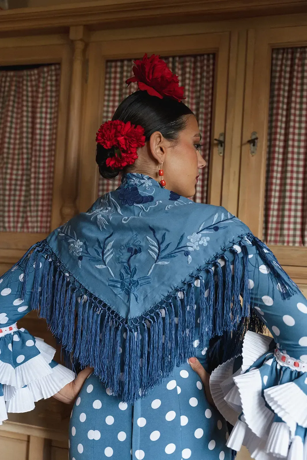 Mujer con un vestido de lunares azules, chal floral y adornos para el cabello posa frente a un gabinete de madera.