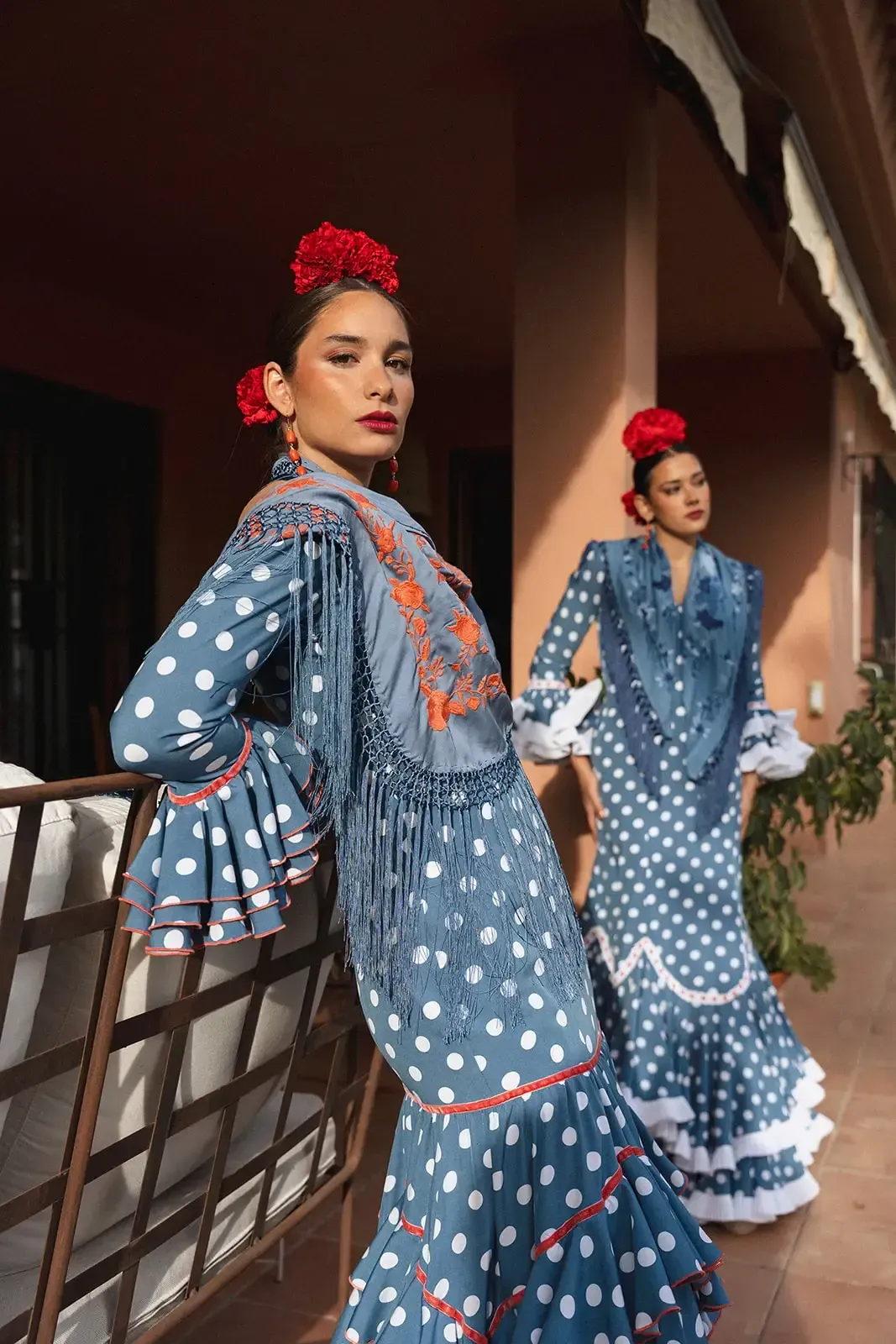 Dos mujeres con trajes de flamenca de lunares y tocados de flores rojas, posando en un balcón.