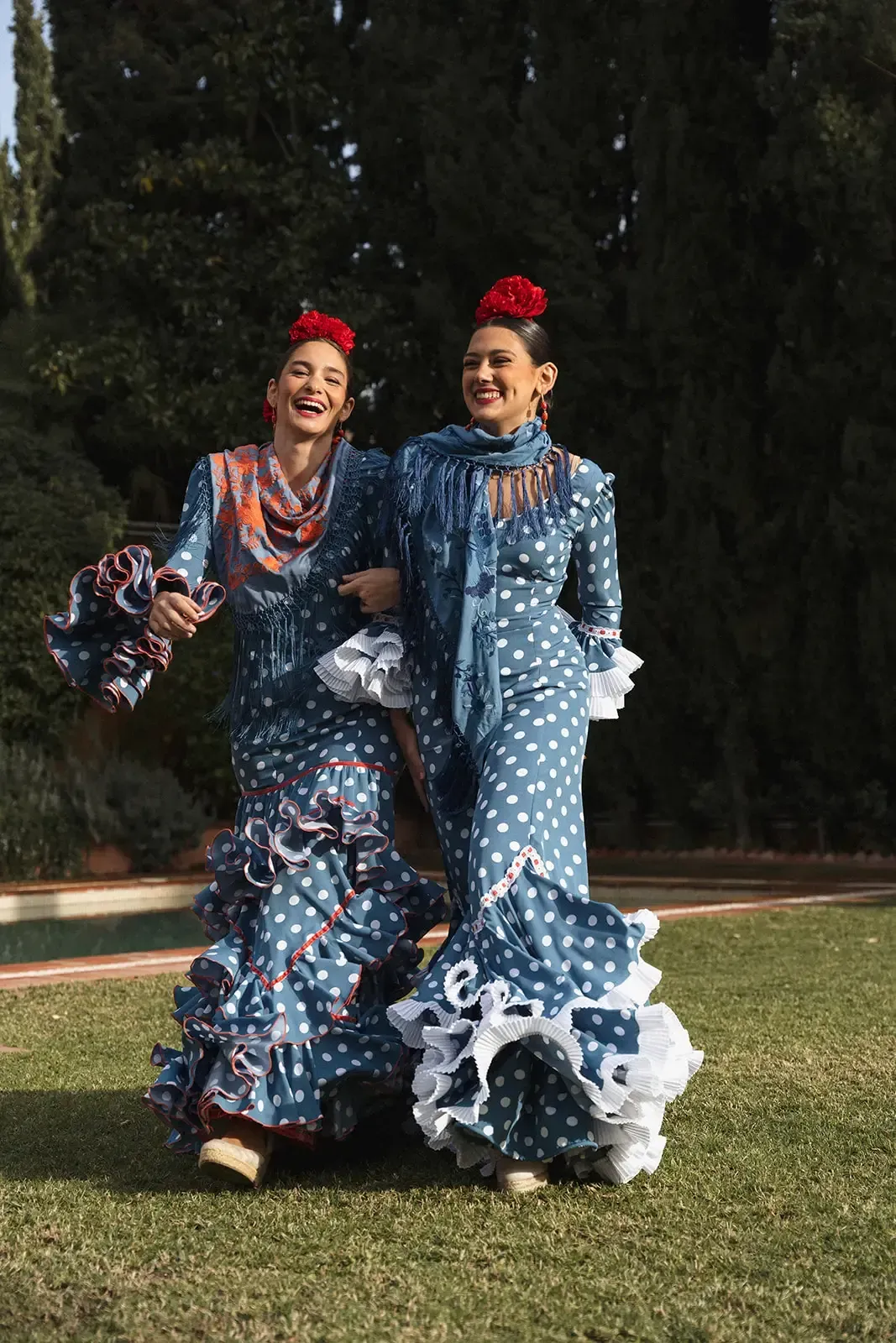 Dos mujeres con trajes de flamenca de lunares azules caminan del brazo por un parque. Llevan flores rojas en el pelo y sonríen.