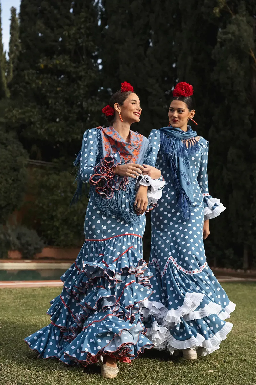 Dos mujeres con vestidos de flamenca de lunares azules, faldas con volantes y flores rojas en el pelo, al aire libre.
