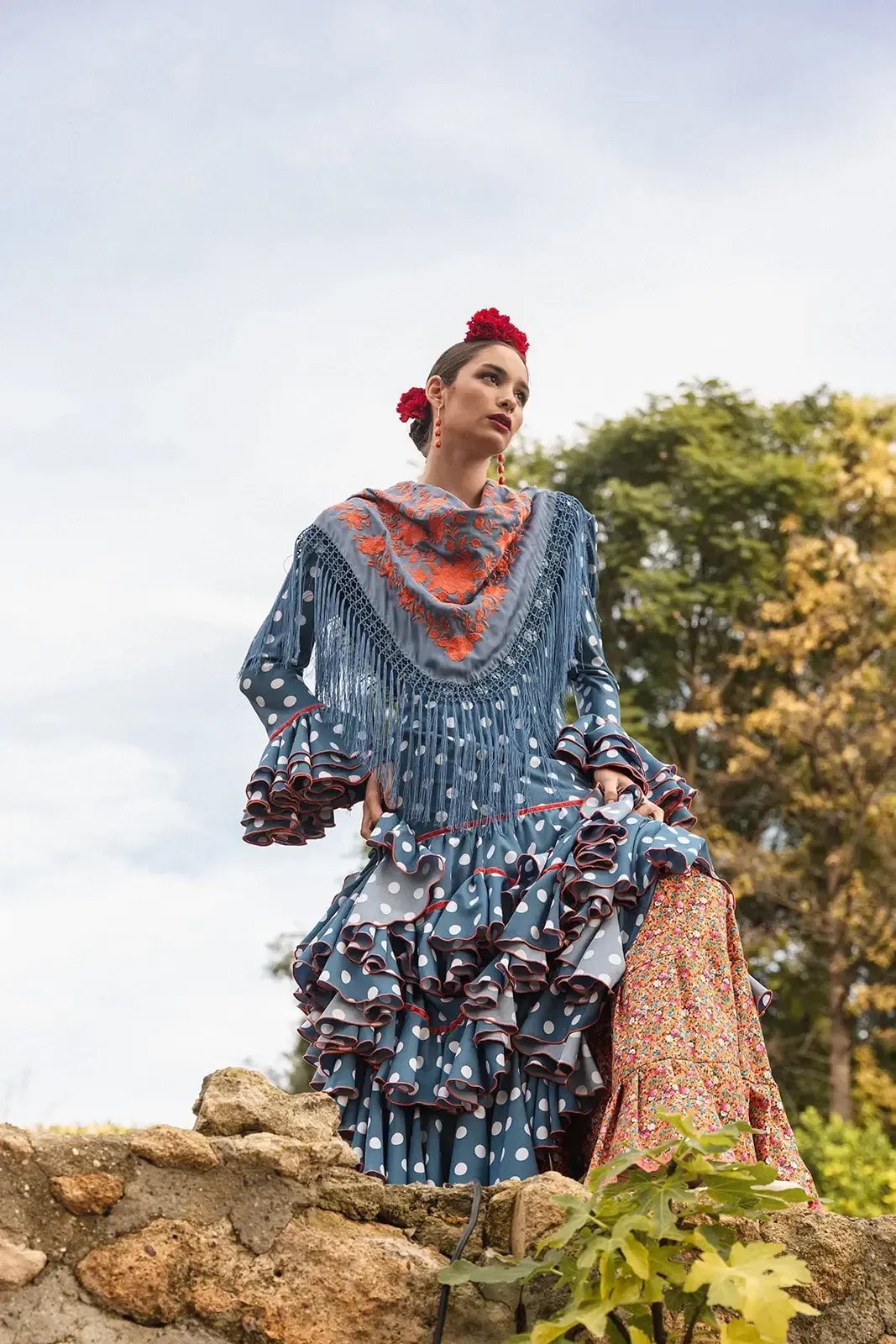 Mujer con traje de flamenca azul con lunares rojos, posando al aire libre con flores rojas en el pelo.