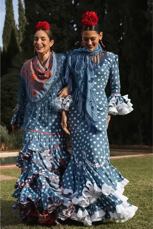 Dos mujeres con vestidos de lunares azules con volantes, tomadas del brazo, sonriendo al aire libre, flores rojas en el pelo.