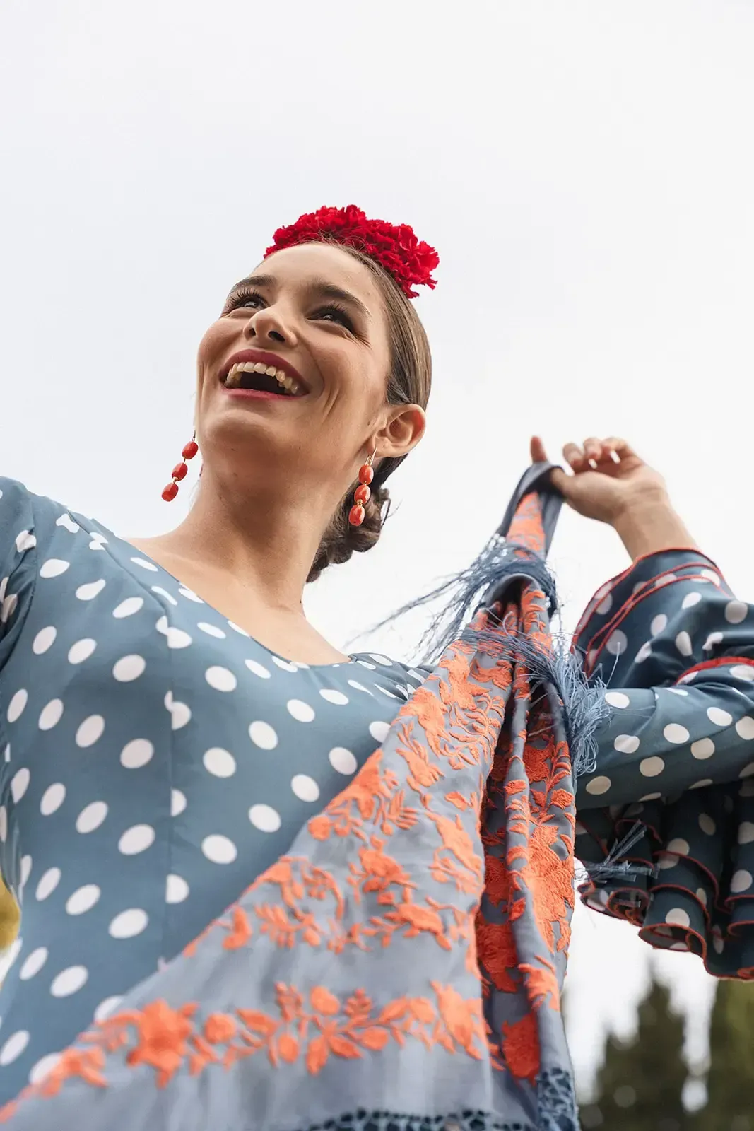 Una mujer sonríe, bailando, vistiendo un vestido de lunares con un chal con flecos naranjas y una flor en el pelo.
