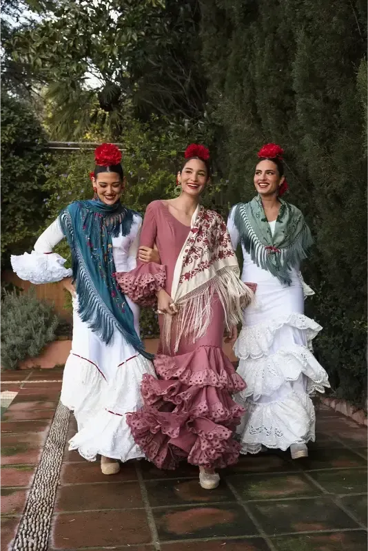 Tres mujeres con trajes de flamenca posan al aire libre, luciendo mantones y flores rojas en el pelo.