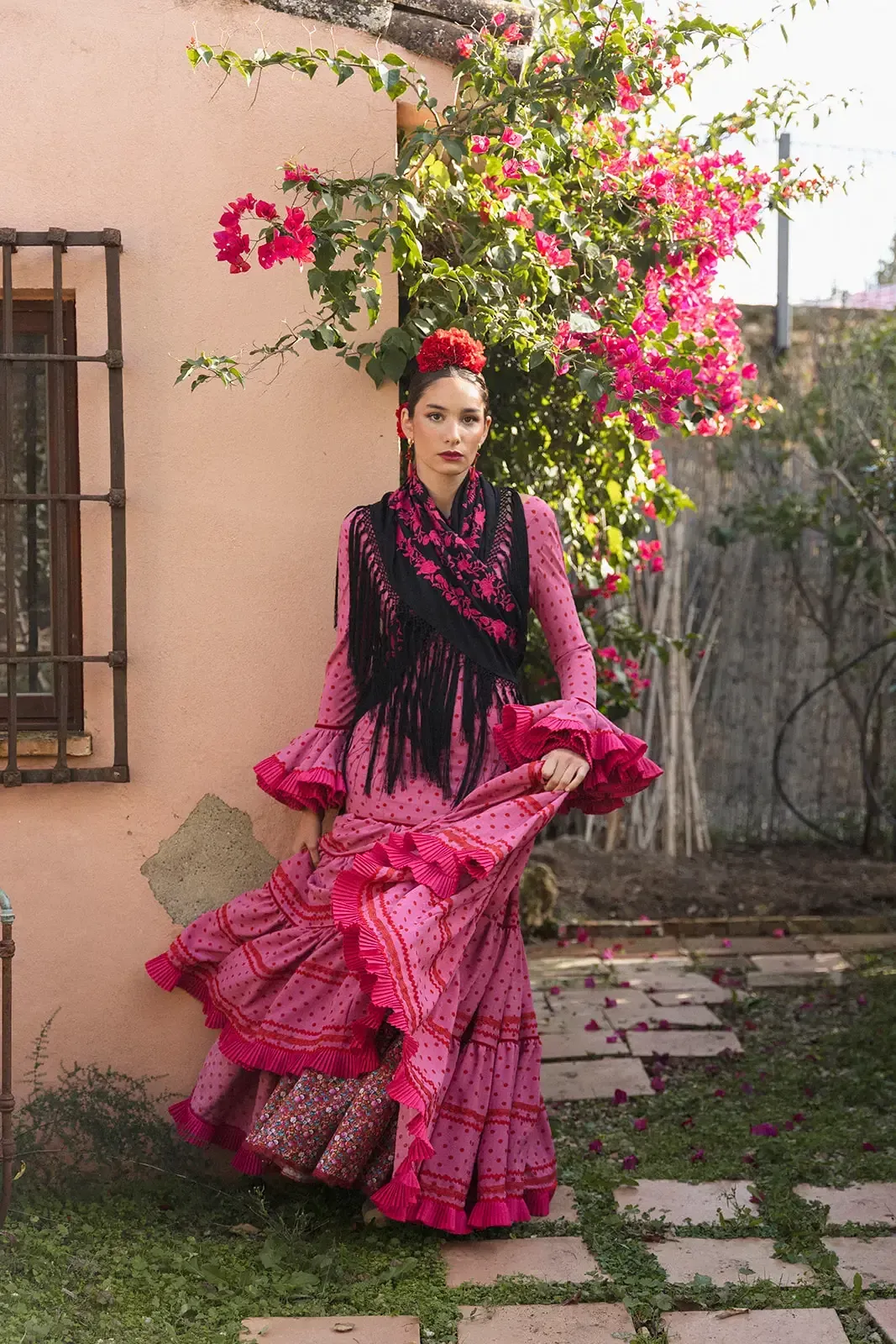 Mujer con traje de flamenca rosa, mantón y flor en el pelo, cerca de un edificio con buganvillas rosas.