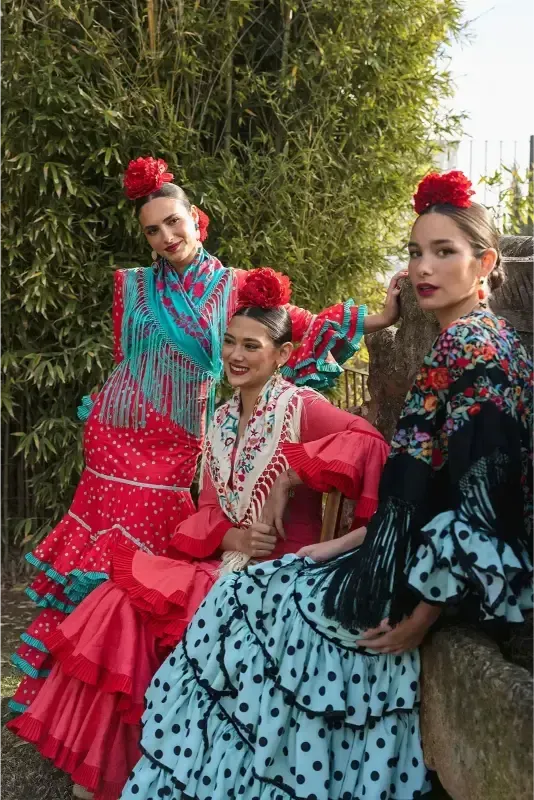 Tres mujeres con coloridos trajes de flamenca y flores en el pelo, al aire libre.