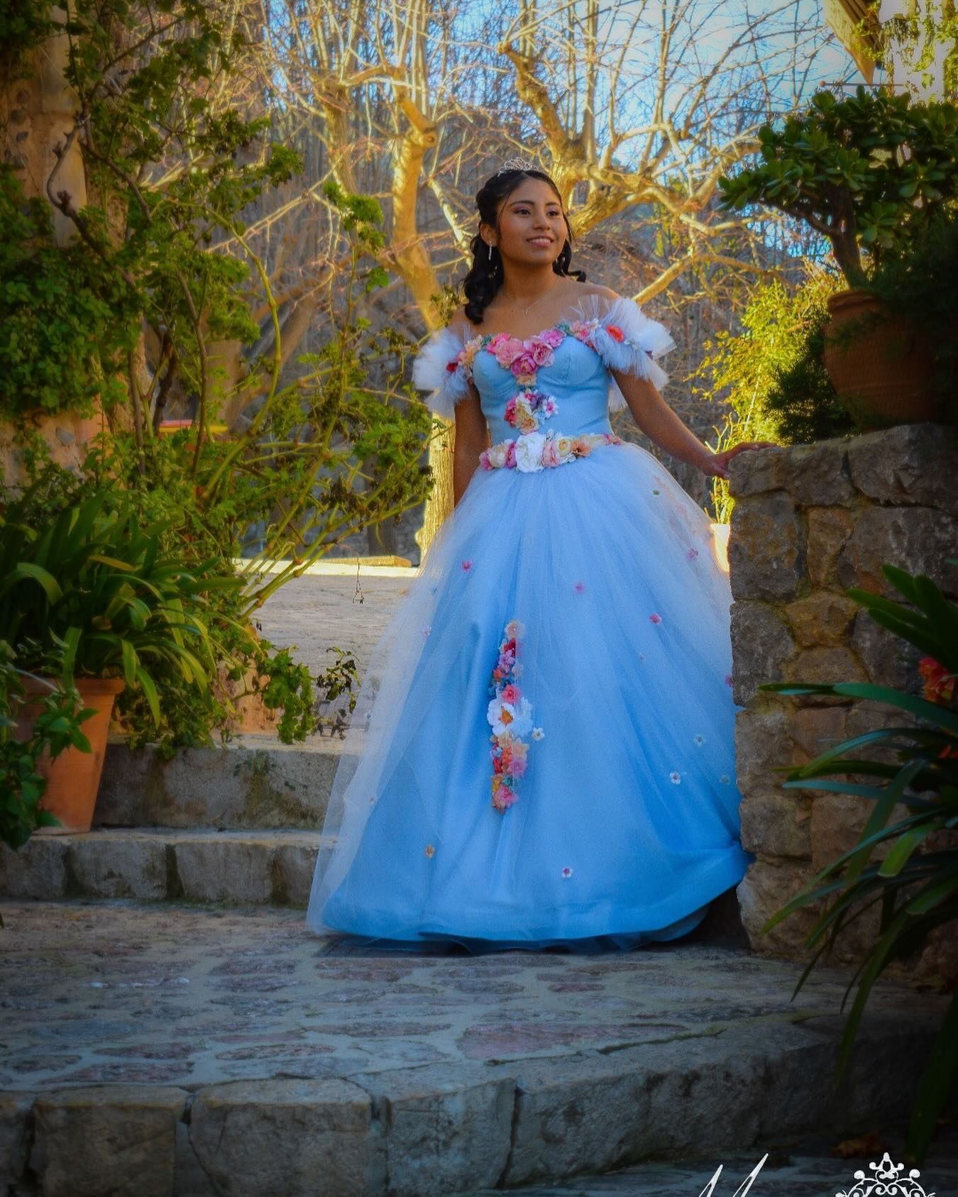 Una mujer con un vestido azul claro posa en unos escalones de piedra de un jardín, sonriendo.