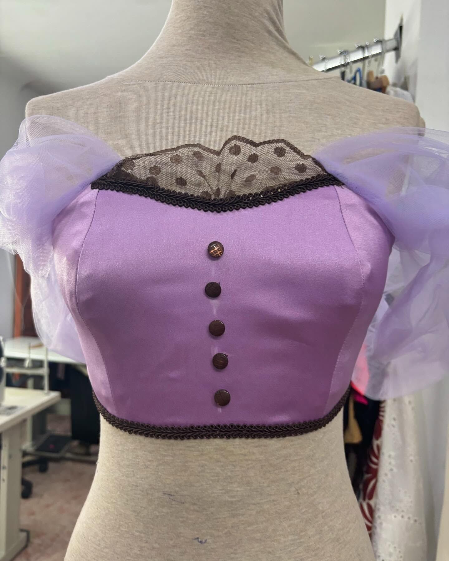 Corsé de satén lavanda con ribete y botones marrones, combinado con mangas violetas transparentes sobre un maniquí.