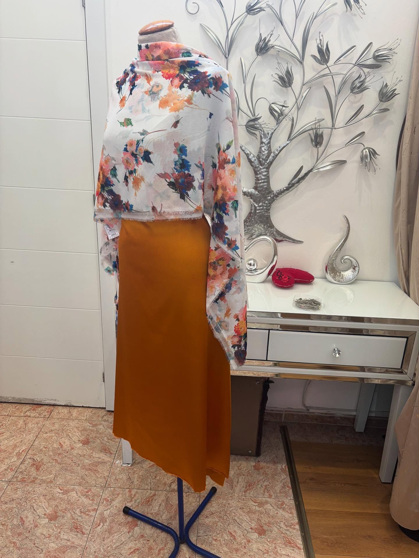 Maniquí con vestido naranja y chal floral en una habitación con una mesa decorativa y un árbol.